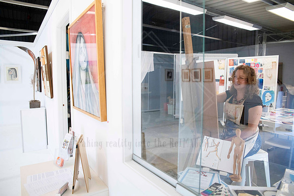 Artist waving from inside gallery window, surrounded by framed artworks, captured for Artwalk event visuals.