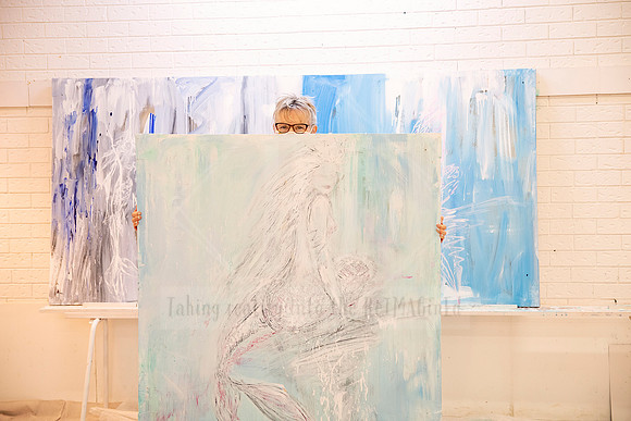 Artist peeking over a large pastel-toned canvas with abstract female figures, standing against matching artwork.