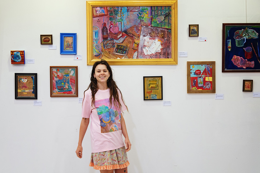 Lucy Juhnke stands smiling in front of a wall of her paintings at Facts and Figurines. She wears a pink t-shirt featuring her own artwork, surrounded by small and large pieces in bright frames. The image celebrates the joy of creation and the power of art to fill space with colour and personality.