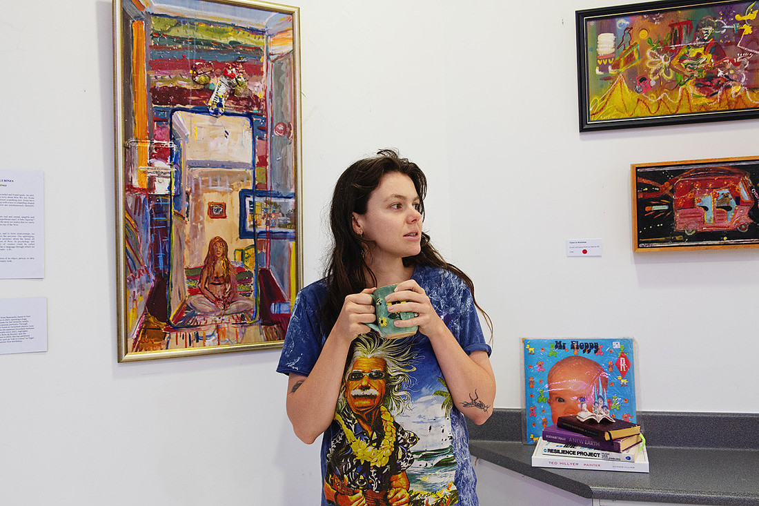 Artist Lucy Juhnke pauses with a ceramic cup in hand beside her expressive paintings during her Facts and Figurines exhibition. The portrait captures her natural presence among colourful canvases and creative props, reflecting the warmth and authenticity of her artistic process.