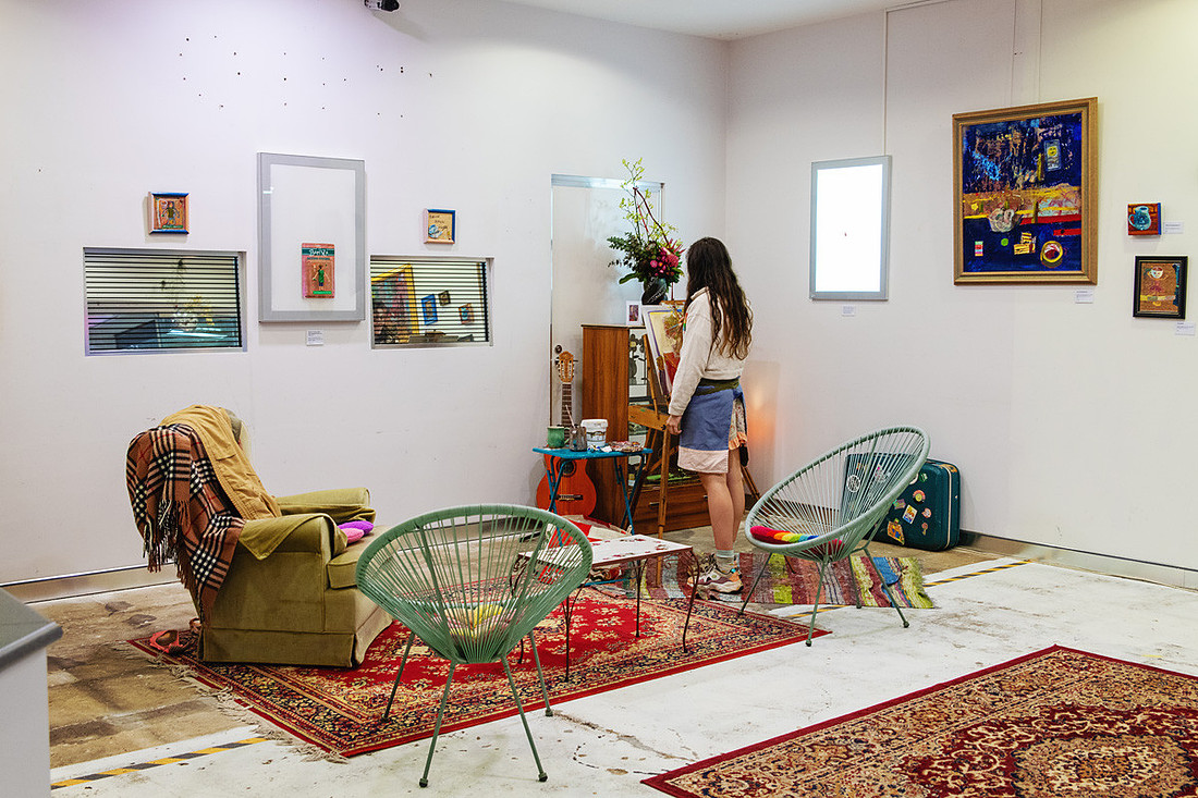 Wide view of Lucy Juhnke’s exhibition space at the Port Central Make/Space in Port Macquarie, showing her at the easel surrounded by mid-century furniture, rugs, and colourful artworks. The scene feels homely and conversational — blending living space and gallery to invite connection and artistic dialogue.