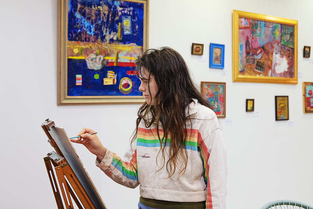 Artist Lucy Juhnke paints at her easel during the Facts and Figurines exhibition in Port Macquarie. Behind her, a wall of vivid, textured artworks glows with layers of blue, pink, and gold. The photograph captures her focus, technique, and the expressive environment that defines her practice.