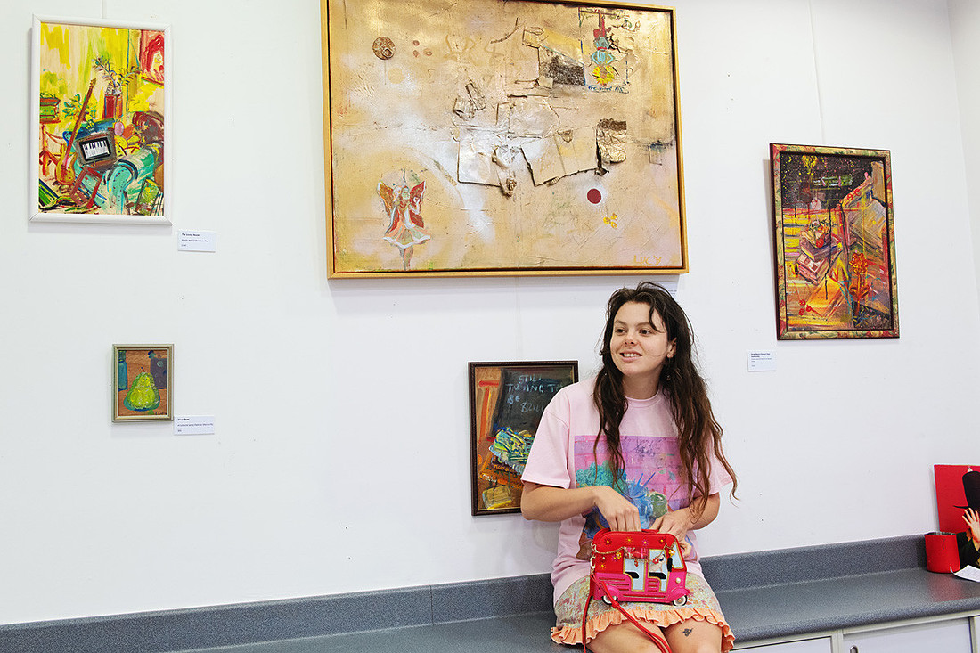 Lucy Juhnke sits casually among her displayed artworks at the Port Central Make/Space. She holds a red handbag while surrounded by her bold, story-filled paintings. The scene shows her ease within her own creative world — a moment of artist and artwork coexisting in full colour.