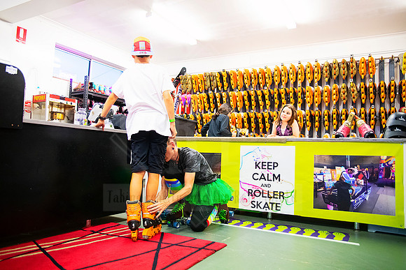 Child being helped to put on skates by a staff member near 'Keep Calm & Roller Skate' sign.