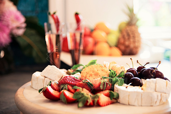 Grazing platter with strawberries, brie, grapes, and crackers against a tropical fruit backdrop.