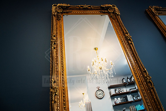 Gold salon mirror reflecting chandelier, shelves, and a touch of moody blue wall paint.