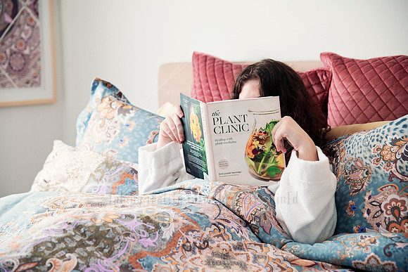 Guest lounging in bed, reading 'Plant Care' book surrounded by colorful linen and cushions.