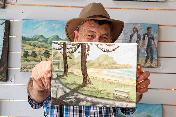 Artist proudly holding landscape painting in front of his displayed works, photographed for Artwalk artist features.