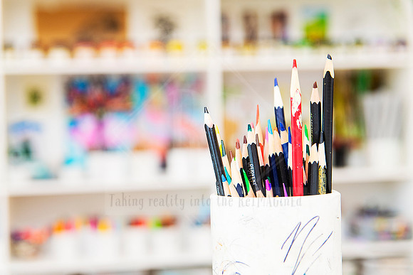 Jar of sharpened coloured pencils in front of a blurred, colourful art shelf.