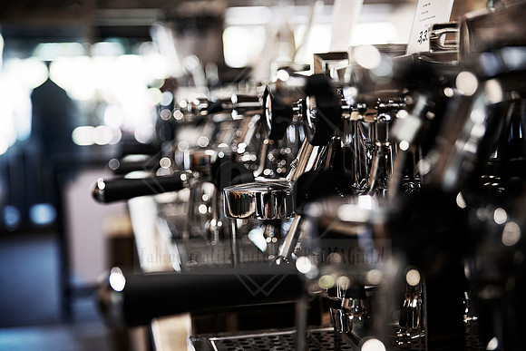Shiny chrome espresso machine group head and handles in soft café lighting.