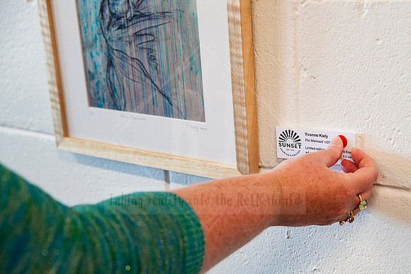 Hand placing exhibition label under a framed artwork, detail capture from gallery prep at Sunset Framing & Gallery.