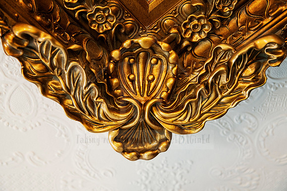Detail of an antique gold corbel with floral and scrollwork carving against white embossed wallpaper.