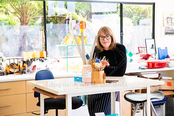 Glass artist seated in a bright studio workspace with colourful supplies, photographed for Artwalk content series.
