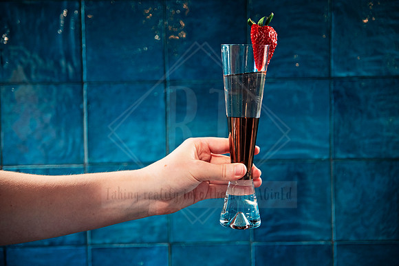 Hand holding a champagne flute with a fresh strawberry garnish in front of teal tiles.