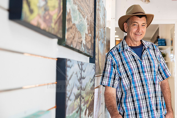 Smiling artist standing beside his work, wall of painted landscapes in the background, part of Artwalk’s artist profiles.