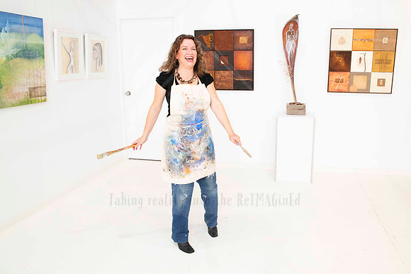 Artist in paint-splattered apron smiling and gesturing in white gallery space, photographed for Artwalk celebration content.