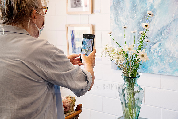 Woman with short grey hair photographing her artwork on a white wall using a mobile phone.