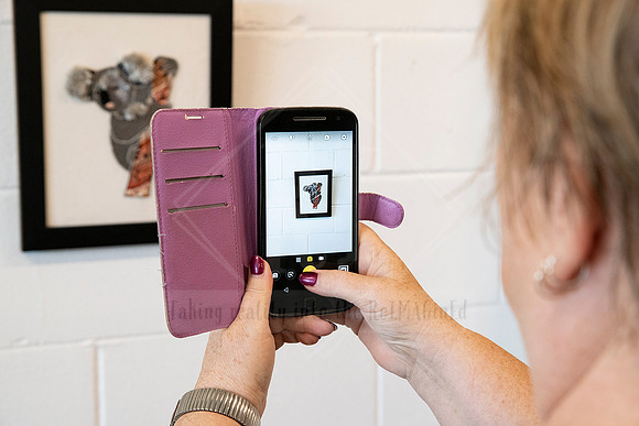 Woman photographing framed art on a white wall using her phone, captured inside Sunset Framing & Gallery.