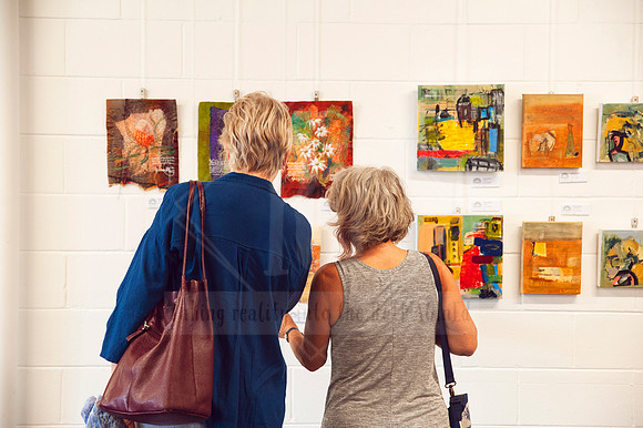 Two women viewing small artworks at Sunset Framing & Gallery, photographed for community engagement and arts marketing content.