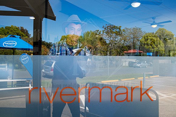 Reflection shot of “Rivermark” signage with artist’s face visible in glass, blending place and presence for Artwalk feature.