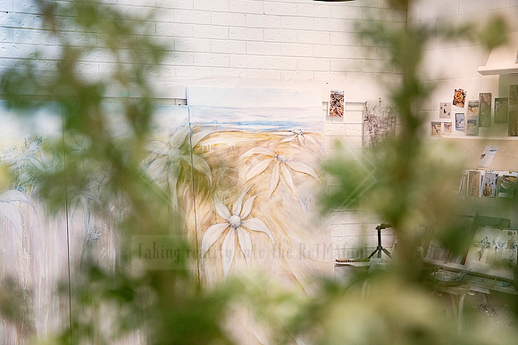 Gallery wall partially obscured by green foliage, with artworks and prints on display.
