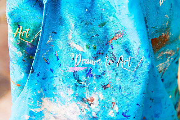 Close-up of a blue 'Drawn To Art' apron, marked with multicolour paint splatters.