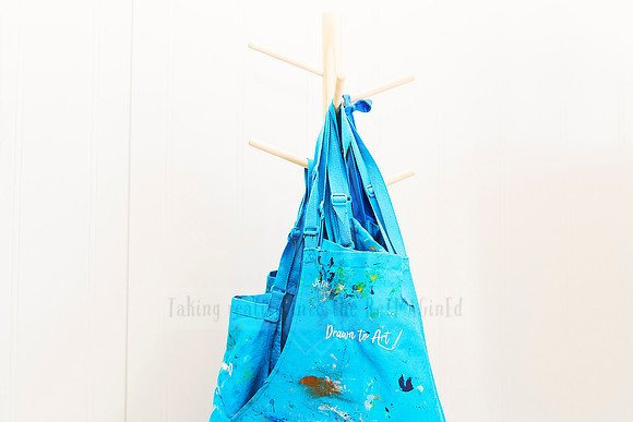 Blue paint-splattered aprons hanging from a wall peg, branded with 'Drawn To Art'.