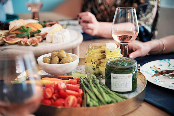 Hands reaching over a colourful grazing platter with olives, dips, asparagus, and wine.
