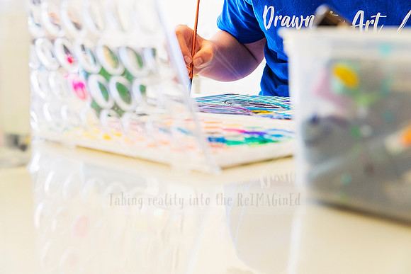 Artist mixing paint on a palette while painting, with colourful materials in the foreground.