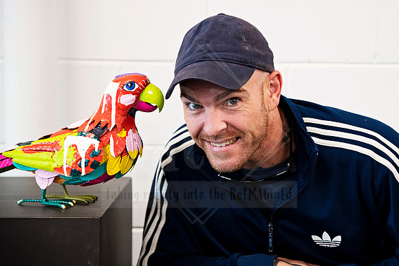 Smiling man leaning near a brightly painted bird sculpture, photographed for Sunset Framing & Gallery content.