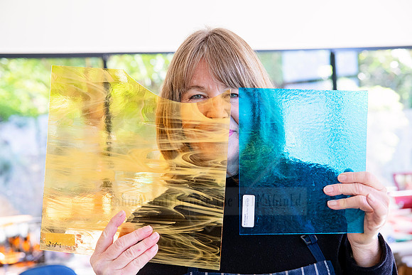 Glass artist peeking from behind vibrant blue and gold glass panels, captured for Artwalk’s artist spotlight series.