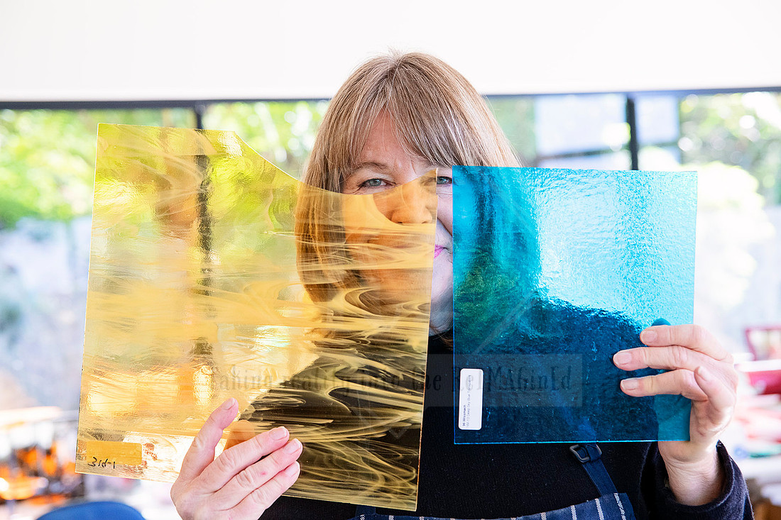 Glass artist peeking from behind vibrant blue and gold glass panels, captured for Artwalk’s artist spotlight series.