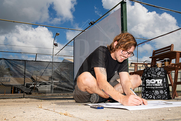 Artist crouching in front of mural space sketching design onto canvas outdoors, captured during Artwalk street art prep.